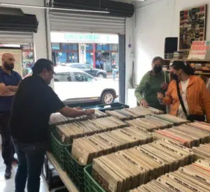 Four people browse through rows of vinyl records in a record store, with large windows and a street visible outside.